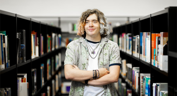 A young male with wavy multicoloured hair is standing with his arms crossed, smiling in the library