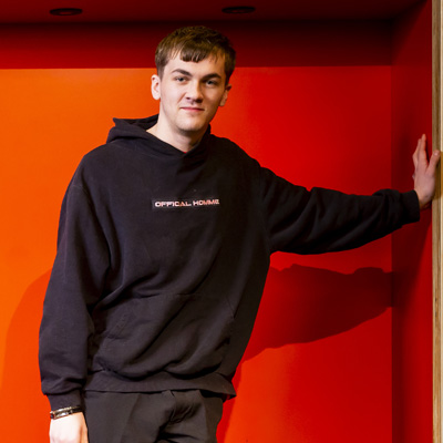 A young male student wearing black casual wear is standing casually in front of a red background