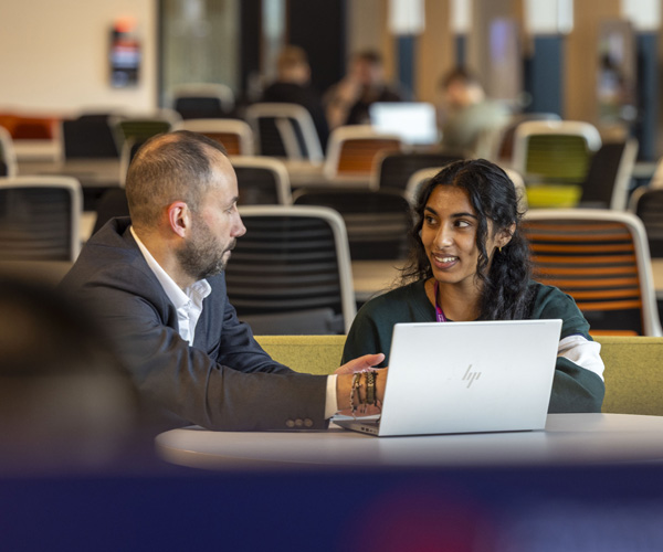 A female student with dark hair and a male lecturer dressed smartly are sitting with a laptop in a modern open collaborative space