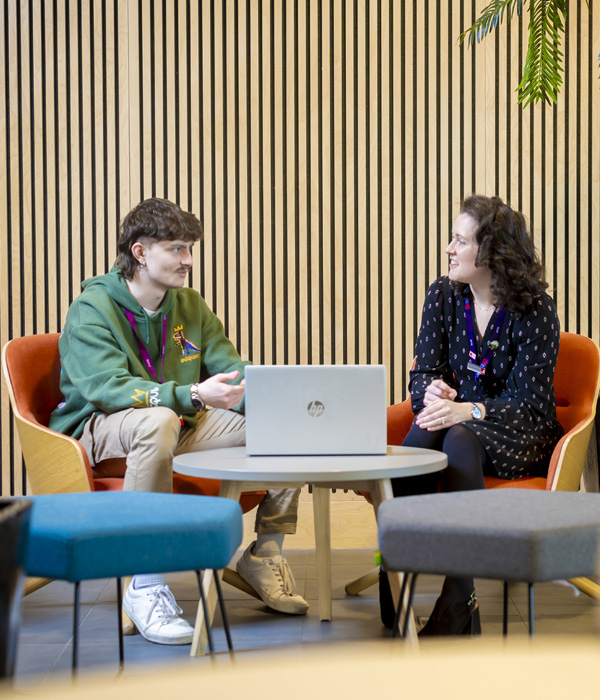 A young male student wearing a green hoody is sitting with a female academic, they are chatting with a laptop