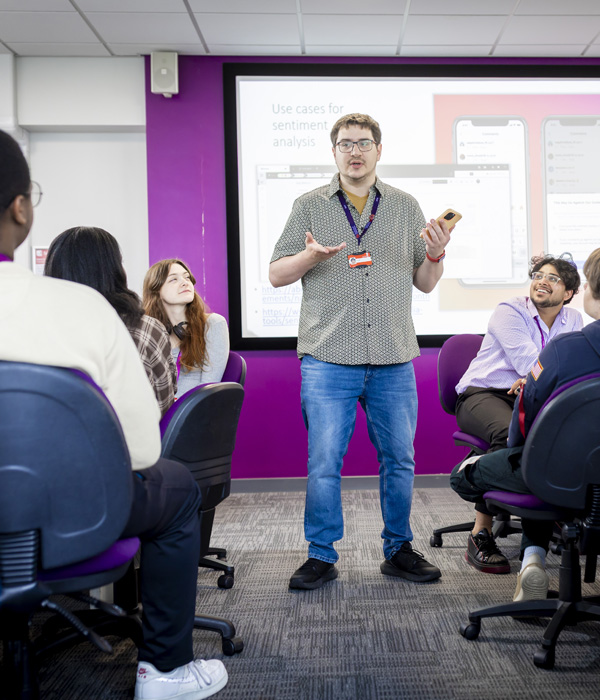 Students are in the computer seminar room, there is a male teacher standing at the front presenting to the students