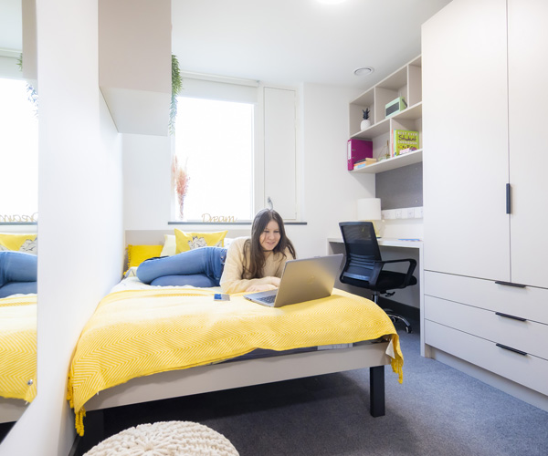A young female student is relaxing on her bed using her laptop in a modern student room