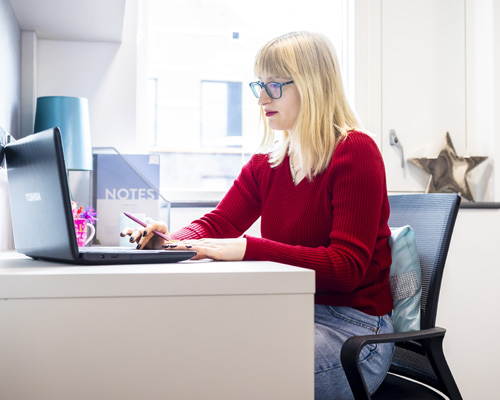 A female with blonde hair wearing a red jumper is using her laptop in modern student accommodation