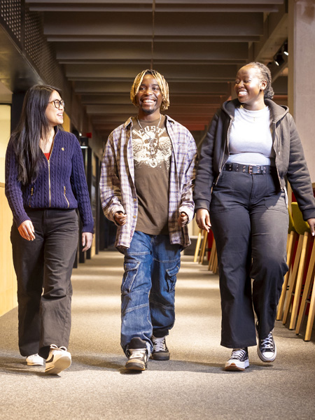Three young students wearing casual clothing are walking and smiling together through a modern study space
