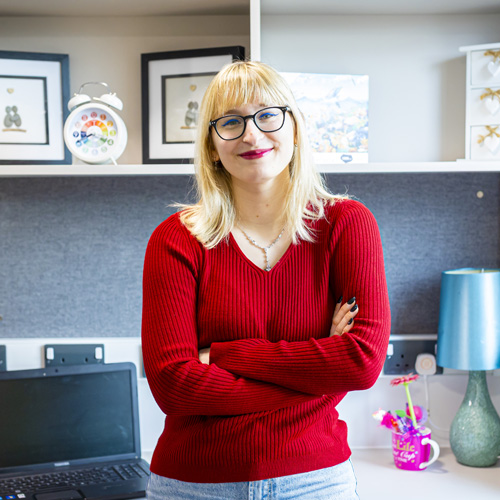 A female with blonde hair and glasses is smiling and standing confidently infront of her desk in student accommodation
