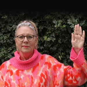 Woman in a bright pink and orange jumper raises one hand against a dark leafy background.