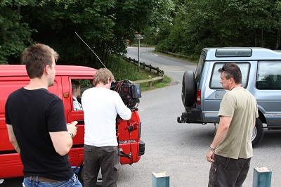Small film crew records a scene beside red and blue vans on a quiet roadside.