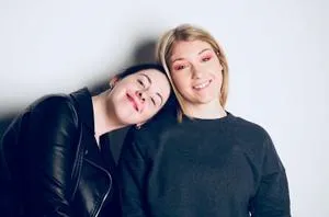 Two young women pose together, one resting her head on the other's shoulder.