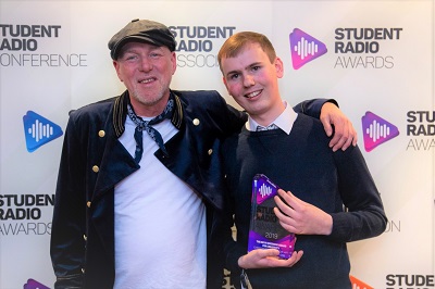 Two men pose at the Student Radio Awards, with the younger man holding a trophy.