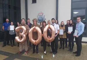 Group holds certificates and large rose-gold balloons forming the number 300.