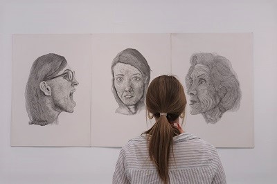 Visitor views three large pencil portraits displayed on a white gallery wall.