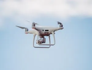 White quadcopter drone hovers against a pale blue, lightly clouded sky.