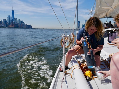 Scientist leans over a sailboat to collect a water sample, with a city skyline behind.