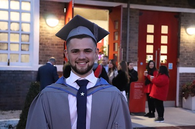 Smiling graduate in cap and gown stands outside a university entrance.