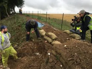 Two men looking into a large hole that has been dug into the ground, while a man and woman look on with a video camera