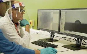 Two laboratory workers in face shields and gloves inspect data on computer monitors.