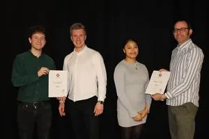 Four people pose on a dark stage, with two students holding certificates.