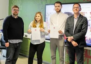 Two students hold certificates while standing between two staff members in a classroom.