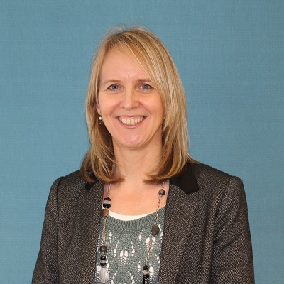 Smiling blonde woman in a dark blazer poses against a muted blue background.