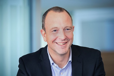 Smiling man in a dark suit and pale blue shirt poses against a softly blurred office background.
