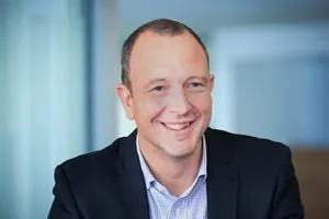 Smiling man in a dark suit and pale blue shirt poses against a softly blurred office background.