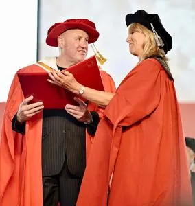 Man and woman in red academic robes exchange a diploma during a graduation ceremony.