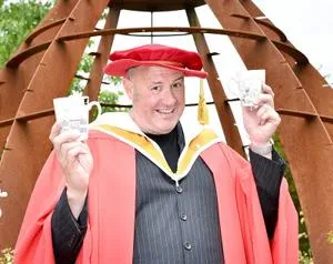 Man in red academic robes holds two small ceramic pieces in front of a rust-coloured sculpture.