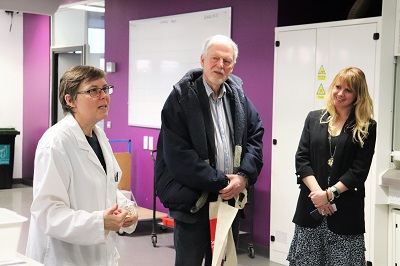 Woman in a white lab coat speaks with two visitors during a tour of a science laboratory.