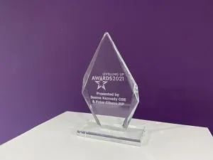 Clear glass Levelling Up Awards 2021 trophy displayed on a white table against a purple wall.