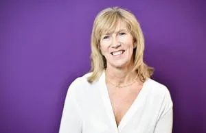 Smiling blonde woman in a white blouse poses against a purple studio background.
