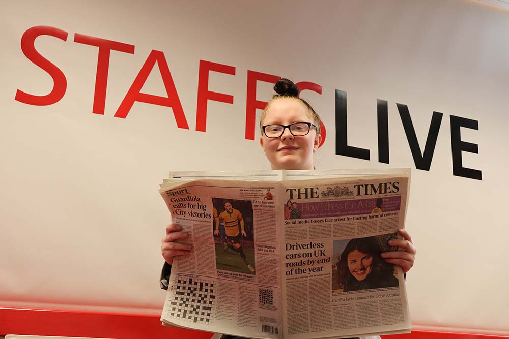 Woman holds an open newspaper beneath large red and black 'STAFFSLIVE' lettering.