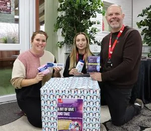 Two people kneel beside a large donation box while holding packaged hygiene products.
