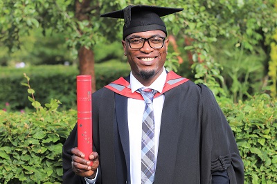 Smiling graduate in cap and gown holds a red diploma tube outdoors.