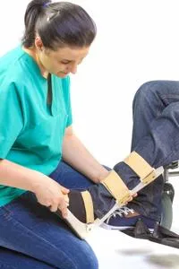 Clinician in green scrubs fits a wooden and fabric orthotic support around a seated person's leg.