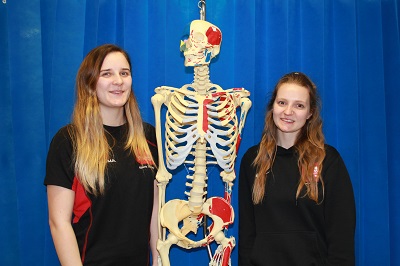 Two smiling women stand on either side of a human skeleton model.
