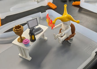 A Playmobil figure sat at a desk holding flames and wearing a jester's hat