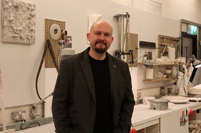 Bald, bearded man in a dark jacket stands inside a ceramics workshop.