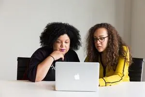 Two women sit at a laptop, with one pointing at the screen as they review results.