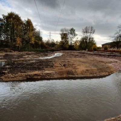 Reworked riverbank with bare soil, shallow water and leafless trees under a cloudy sky.