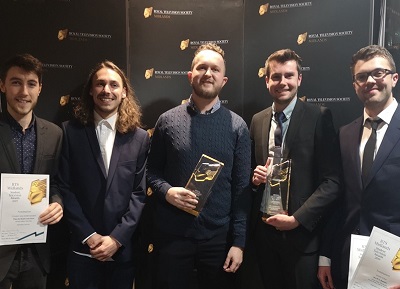 Five men pose at an awards backdrop holding trophies and certificates.