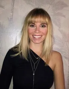 Smiling blonde woman in a black one-shoulder top poses against a pale patterned wall.