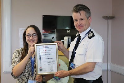 Smiling woman and uniformed police officer hold a framed certificate in an office.