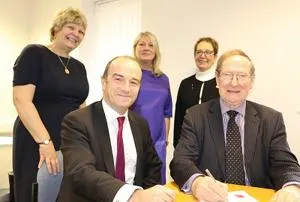 Two men sign a document at a table while three women stand behind them.