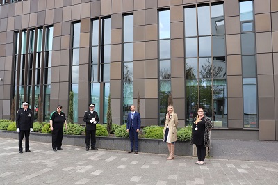Six people stand spaced apart outside a modern brown-clad university building.