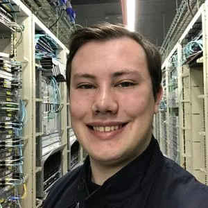 Smiling young man takes a selfie between rows of equipment in a server room.