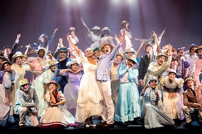 Large musical theatre cast in colourful costumes celebrates together on stage.