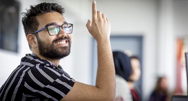 A male student wearing glasses a dark striped shirt is smiling in the classroom