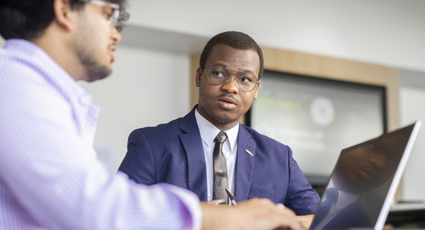 A young male student wearing a blue blazer shirt and tie is sitting at a desk talking to a smartly dressed man using a laptop