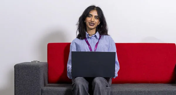 A female student with dark medium length hair wearing a blue shirt is smiling working on her laptop