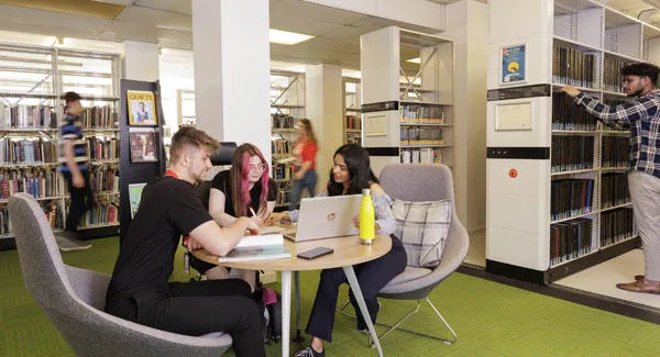 A group of students are in the library sitting together at a table with laptops and books